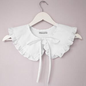 May include: White cotton collar with a ruffled edge and a bow tie. The collar is on a white hanger and has a label that says "MokushArt".