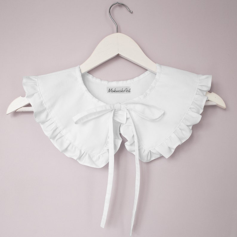 May include: White cotton collar with a ruffled edge and a bow tie. The collar is on a white hanger and has a label that says "MokushArt".