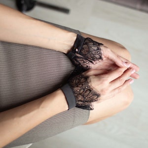 May include: Black lace wrist cuffs with a scalloped edge. The cuffs are attached to a black band with a gold-colored button closure. The cuffs are worn on the wrists.