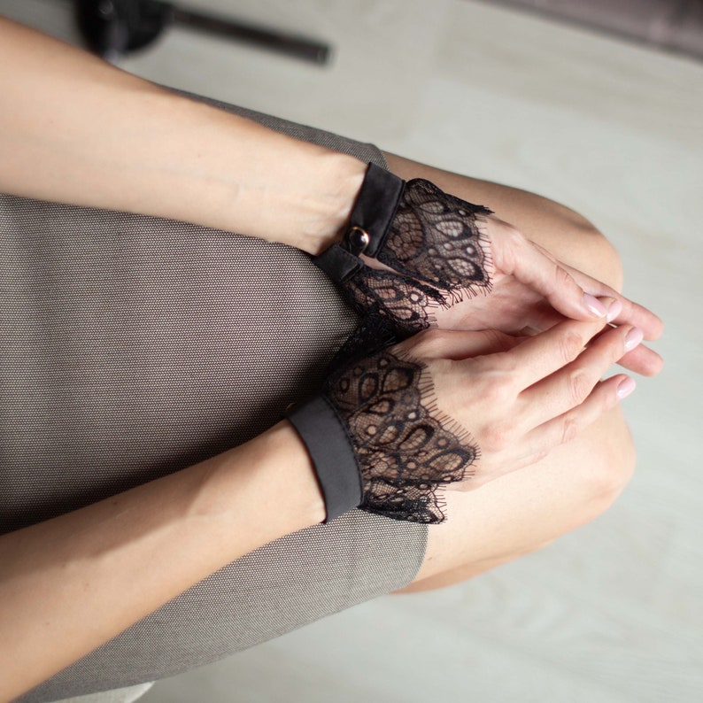 May include: Black lace wrist cuffs with a scalloped edge. The cuffs are attached to a black band with a gold-colored button closure. The cuffs are worn on the wrists.