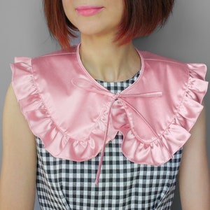May include: A pink satin collar with ruffled edges and a bow tie closure. The collar is a soft pink color and appears to be made of a smooth, shiny fabric. The collar is worn over a black and white checkered top.