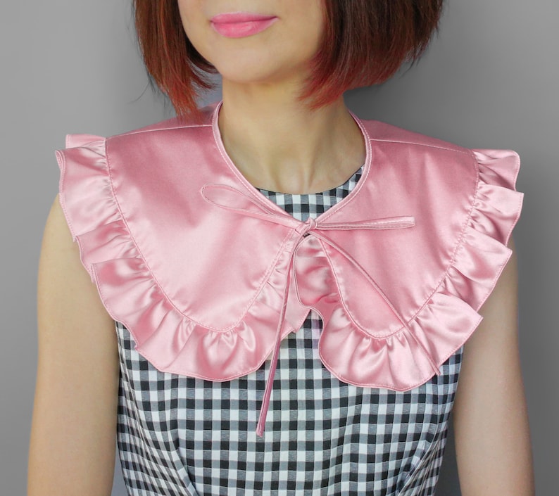 May include: A pink satin collar with ruffled edges and a bow tie closure. The collar is a soft pink color and appears to be made of a smooth, shiny fabric. The collar is worn over a black and white checkered top.