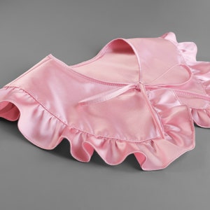 May include: A light pink satin collar with ruffled edges and a tie closure. The collar is designed to be worn around the neck and adds a touch of elegance. The fabric has a slight sheen.