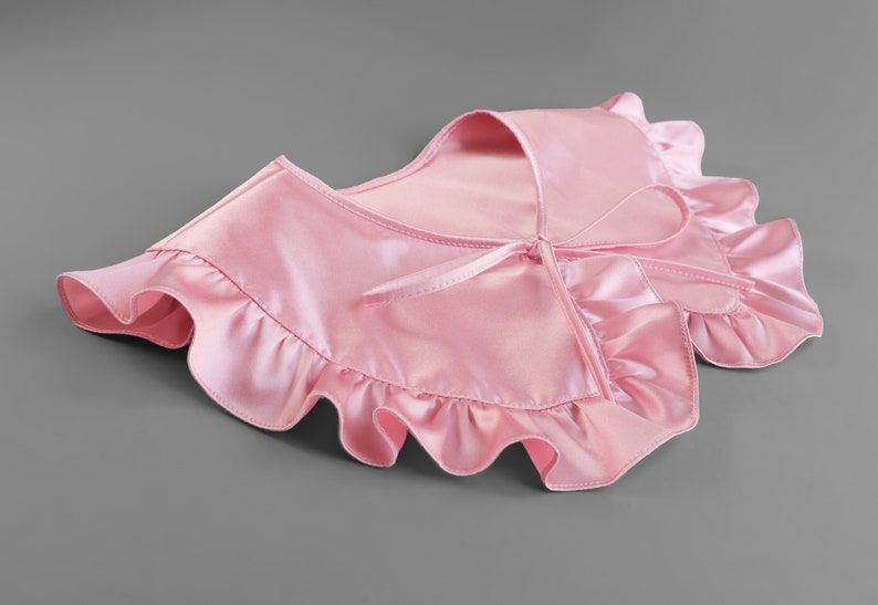 May include: A light pink satin collar with ruffled edges and a tie closure. The collar is designed to be worn around the neck and adds a touch of elegance. The fabric has a slight sheen.