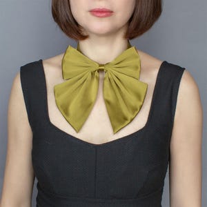 May include: A large, olive green satin bow tie worn around the neck. The bow tie is tied in a large, loose knot.