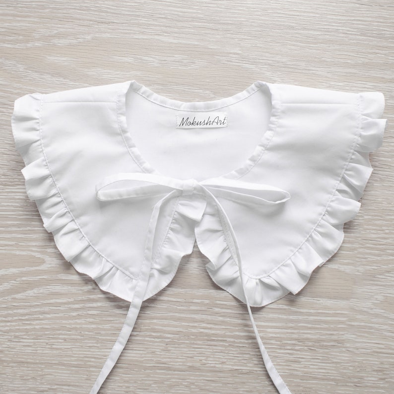 May include: White cotton peter pan collar with a ruffled edge and a bow tie. The collar has a label that says "MokushiAri".