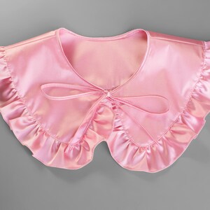May include: A light pink satin collar with a ruffled edge and a bow tie closure. The collar is a soft, feminine accessory, perfect for adding a touch of elegance to any outfit. The collar is a solid color.