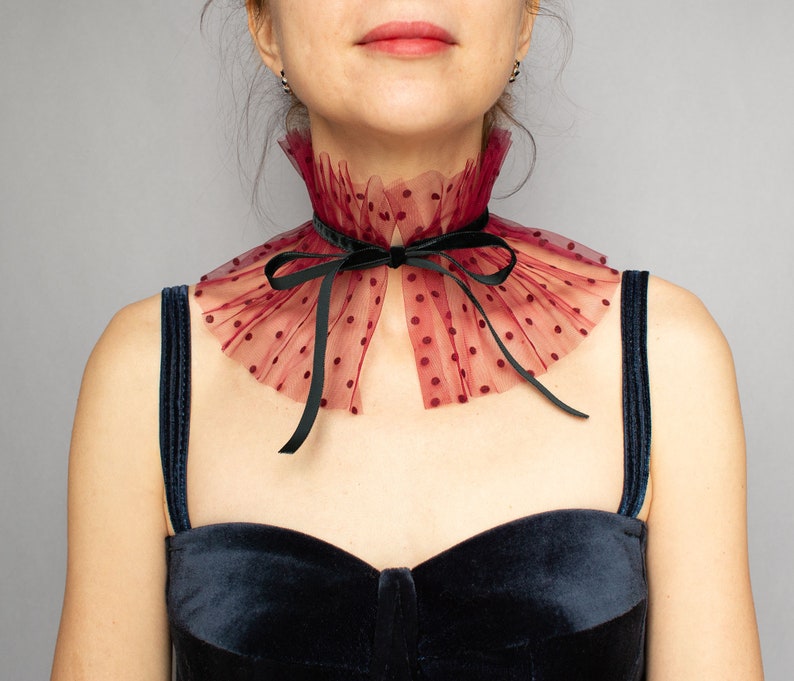 Puede incluir: Un collar de encaje burdeos con lunares negros y un lazo de terciopelo negro. El collar lo lleva una persona con un top de terciopelo negro.