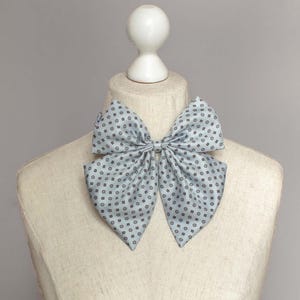 May include: A light blue bow tie with a small white and gray geometric pattern. The bow tie is tied and displayed on a white mannequin.