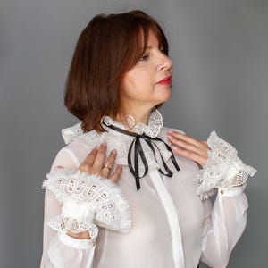 May include: A white sheer long-sleeve blouse featuring a high ruffled lace collar with a thin black ribbon tie at the neck. The blouse includes matching wide lace cuffs with a fringed edge that extend over the wrists, creating a layered appearance.