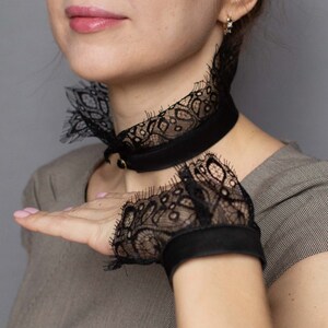 May include: Black lace choker necklace and matching fingerless gloves. The choker features a black satin band with a delicate lace overlay and a small gold-colored clasp. The gloves have a similar lace design and satin trim.