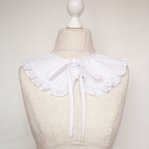 May include: A white ruffled collar with a bow tie. The collar is on a white dress form.
