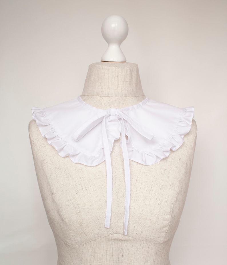 May include: A white ruffled collar with a bow tie. The collar is on a white dress form.
