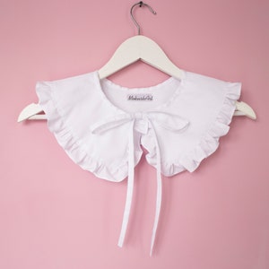 May include: White cotton peter pan collar with ruffled edges and a bow tie. The collar is on a white hanger against a pink background. The label on the collar reads "MakushishArt".