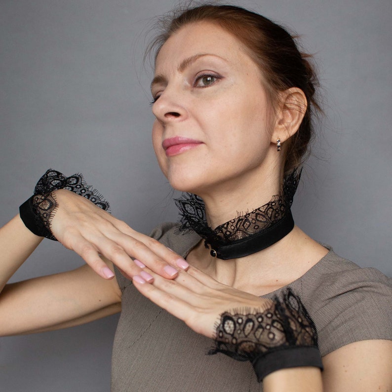 May include: Black lace choker necklace and matching wrist cuffs. The choker is a black band with a delicate lace trim. The wrist cuffs are similar, featuring a black band and intricate lace detailing. The set is designed to be worn together.