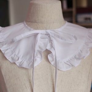 May include: White ruffled collar with a bow tie. The collar is made of a soft, white fabric and has a delicate, ruffled edge. The bow tie is tied at the front of the collar and adds a touch of elegance.