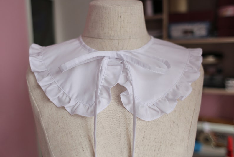 May include: White ruffled collar with a bow tie. The collar is made of a soft, white fabric and has a delicate, ruffled edge. The bow tie is tied at the front of the collar and adds a touch of elegance.