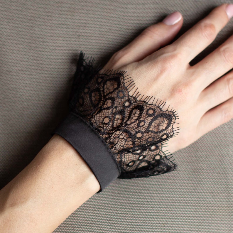 May include: Black lace wrist cuff with a scalloped edge and a solid black band. The delicate lace features an intricate floral pattern. The cuff is worn on the wrist. The background is a solid gray.
