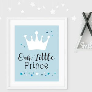 May include: A white framed print with a light blue background. The print features a white crown with a black inscription that reads "Our Little Prince". The background is decorated with small blue and white stars.