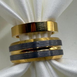 May include: Two gold-colored rings and a ring with a gold band and a dark gray center band are displayed on a white, silky fabric. The rings are simple bands, showcasing a minimalist design.