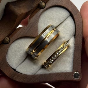 以下が含まれることがあります： 木製のハート型のリングケースには、2つの結婚指輪が入っています。1つは金と銀、もう1つは金に小さな透明な石が付いています。箱は開いており、白い内側が見えます。