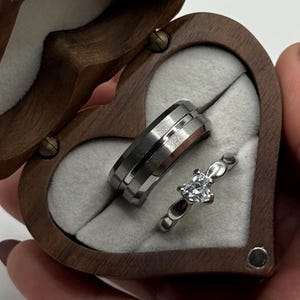 以下が含まれることがあります： ハート型の木製リングボックス。開くと2つの指輪が現れます。1つの指輪は中央にバンドが付いたシルバーで、もう1つは透明な宝石が付いたシルバーバンドです。ボックスは明るい灰色の生地で裏打ちされています。