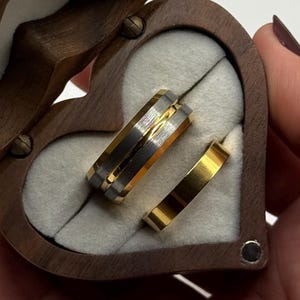 May include: A heart-shaped wooden ring box with a hinged lid, open to reveal two rings. One ring is gold with a silver band, and the other is solid gold. The box is lined with white fabric.