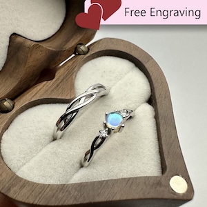 Könnte beinhalten: Zwei silberne Ringe, präsentiert in einer herzförmigen Holzbox mit weißem Innenraum. Ein Ring hat ein verschlungenes Design, der andere einen blauen Edelstein und kleine Diamanten. Auf der Box steht "Free Engraving".