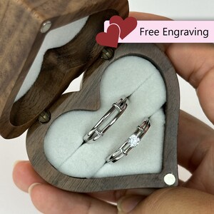 Puede incluir: Una caja de anillos de madera en forma de corazón con un interior de terciopelo, que contiene dos alianzas de plata. La caja está abierta y los anillos son visibles. El texto "Grabado gratuito" está impreso en una etiqueta rosa.