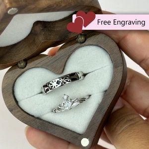 May include: A heart-shaped wooden ring box with a velvet interior, open to reveal two silver rings. The box is made of dark wood and has a hinged lid. The text "Free Engraving" is printed on a pink label.