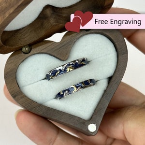 Puede incluir: Dos anillos de plata con esmalte azul y detalles dorados, que presentan un diseño de sol y luna, se muestran en una caja de madera en forma de corazón. La caja está abierta y los anillos son visibles. El texto "Grabado gratuito" está impreso en una etiqueta rosa.