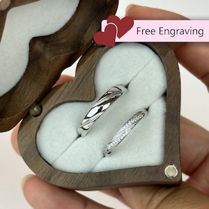 May include: Two silver wedding bands in a heart-shaped wooden box. The box is open and the rings are visible. The text "Free Engraving" is on the box.