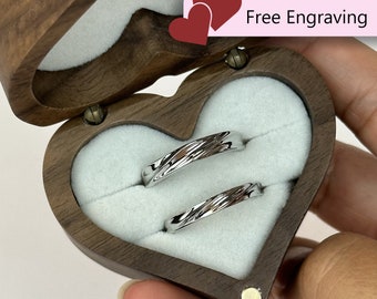 Könnte beinhalten: Zwei silberne Eheringe in einer herzförmigen Holzschachtel mit einem weißen Samt-Innenfutter. Die Schachtel ist geöffnet und die Ringe sind sichtbar. Der Text "Kostenlose Gravur" ist auf der Schachtel aufgedruckt.
