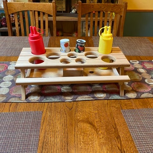 May include: A wooden picnic table condiment holder with nine holes for bottles and jars. The holder has a red and yellow bottle in it, and a small can that says "Pepper".