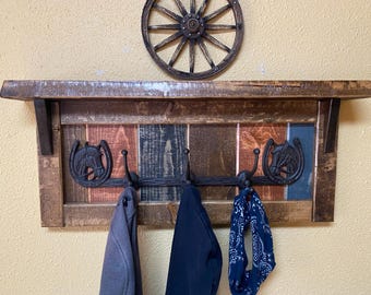 May include: Rustic wooden coat rack with a shelf, featuring a distressed wood finish and a multi-colored design. The rack has five hooks with horse head accents and is decorated with a wagon wheel.