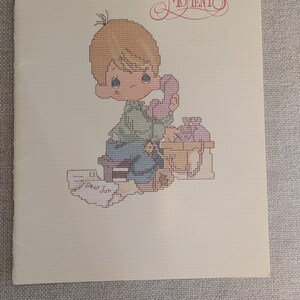 May include: A cross-stitch pattern featuring a Precious Moments design. The image shows a young child with blonde hair holding a pink telephone, with the text "Dear Jon" and "Precious Moments" visible. The color palette includes soft pastels.