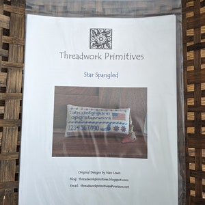 May include: A white paper pattern with the title "Threadwork Primitives" and the subtitle "Star Spangled". The pattern includes a small image of a cross-stitch sampler with the alphabet, numbers, and a small American flag. The text "Original Designs by Nan Lewis" is at the bottom of the page, along with the blog and email address.