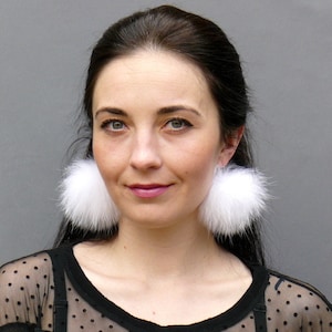 May include: A woman wearing a black sheer top with white polka dots and large white fluffy pom-pom earrings.