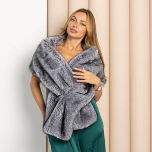 May include: A plush, grey faux fur stole featuring a multi-tonal pattern of dark and light grey fibers. The wrap is draped over the shoulders, showcasing a thick, textured pile and a soft, voluminous appearance that covers the upper torso area.