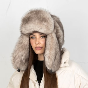 May include: A plush, thick faux fur trapper hat in a light taupe and grey colour palette. The hat features a wide, prominent front brim and long, full ear flaps that extend downwards, providing full coverage with a soft, dense, and textured appearance.