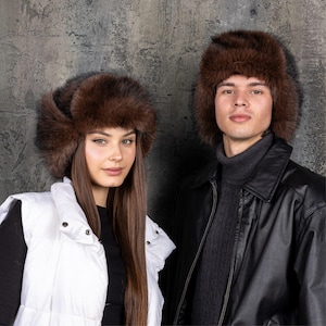 May include: Two brown fur ushanka hats with thick, plush texture and ear flaps. The hats feature a dense, natural-looking fur construction with a rounded crown and wide brim, designed to provide full coverage for the head and ears in cold weather.