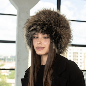 May include: A fluffy, round faux fur hat featuring a mix of dark brown, light brown, and black fibres. The hat has a thick, voluminous texture with long, soft strands that create a full, rounded silhouette around the head, providing a dense appearance.