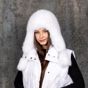 Op de afbeelding: Een witte, pluizige bontmuts met oorflappen en twee hangende pompons, gecombineerd met een wit gewatteerd vest met een hoge kraag en drukknopen. De muts heeft een dikke, dichte textuur en bedekt het hoofd en de oren volledig.