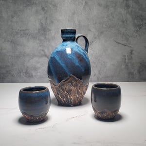 May include: A ceramic drinks set. The jug is blue with brown textured detailing, and has a handle. Two matching small cups are also included. The set has a glossy finish and a mountain-like base.
