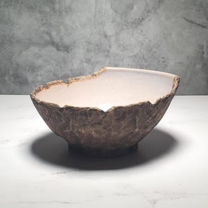 May include: A decorative ceramic bowl with a textured, brown exterior and a smooth, light-coloured interior. The rim has an irregular, organic shape. This handcrafted bowl is suitable for home decor. The bowl's diameter is approximately 25 cm.