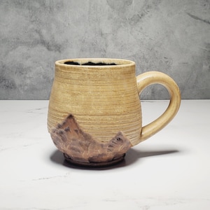 May include: A handmade ceramic mug with a mountain design. The mug is tan with a brown mountain range sculpted on the base. The interior is black, and the handle is rounded. The mug is on a white surface.