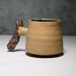 May include: A beige ceramic mug with a natural wood handle. The mug has a rustic, handmade look.