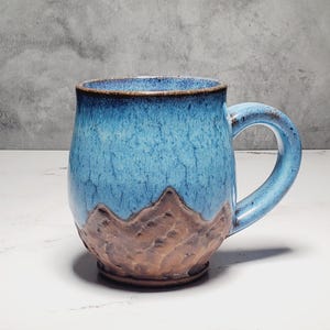 May include: A ceramic mug with a blue and brown mountain design. The mug has a rounded shape with a large handle and a textured surface. The blue glaze transitions to a brown base, creating a mountain-like effect.