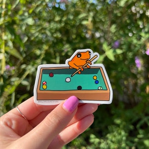 May include: An orange frog wearing a white shirt is playing pool on a green felt table. The frog is holding a pool cue and there are several billiard balls on the table. The table is surrounded by a brown wooden frame. The sticker has the text "@willowdogartstudio" in the bottom left corner.