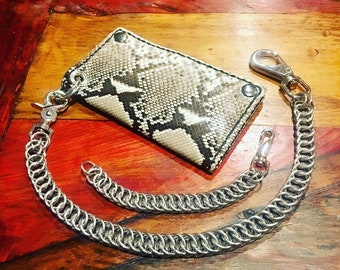 May include: A wallet featuring a snakeskin design in grey and black, accompanied by a silver chain. The chain is constructed from interconnected metal rings and has a clasp at each end. The rectangular wallet has a black border.